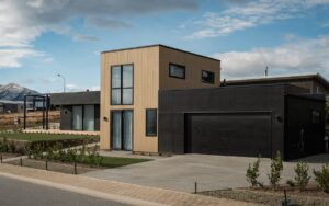 Contemporary home with clean lines and dark cladding, set against a mountain backdrop. A neatly landscaped front yard leads to a deck area, with exposed aggregate concrete bordering the garden.
