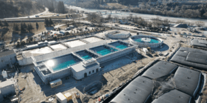 Aerial view of the Shotover Wastewater Treatment Plant upgrade site with multiple tanks and structures under construction, set against the Remarkables mountain range.